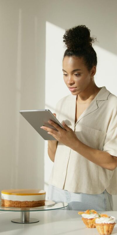 A woman using a tablet in a bright and modern kitchen setting, surrounded by cakes and flowers, depicting a lifestyle scene.