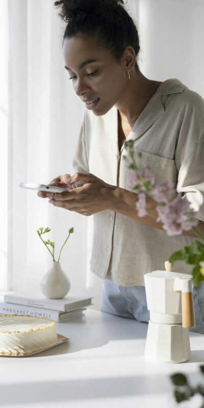 Woman capturing a stylish coffee setup in a sunlit kitchen with flowers and cake.